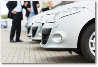 Three people stand behind a row of parked cars on dealership lot.